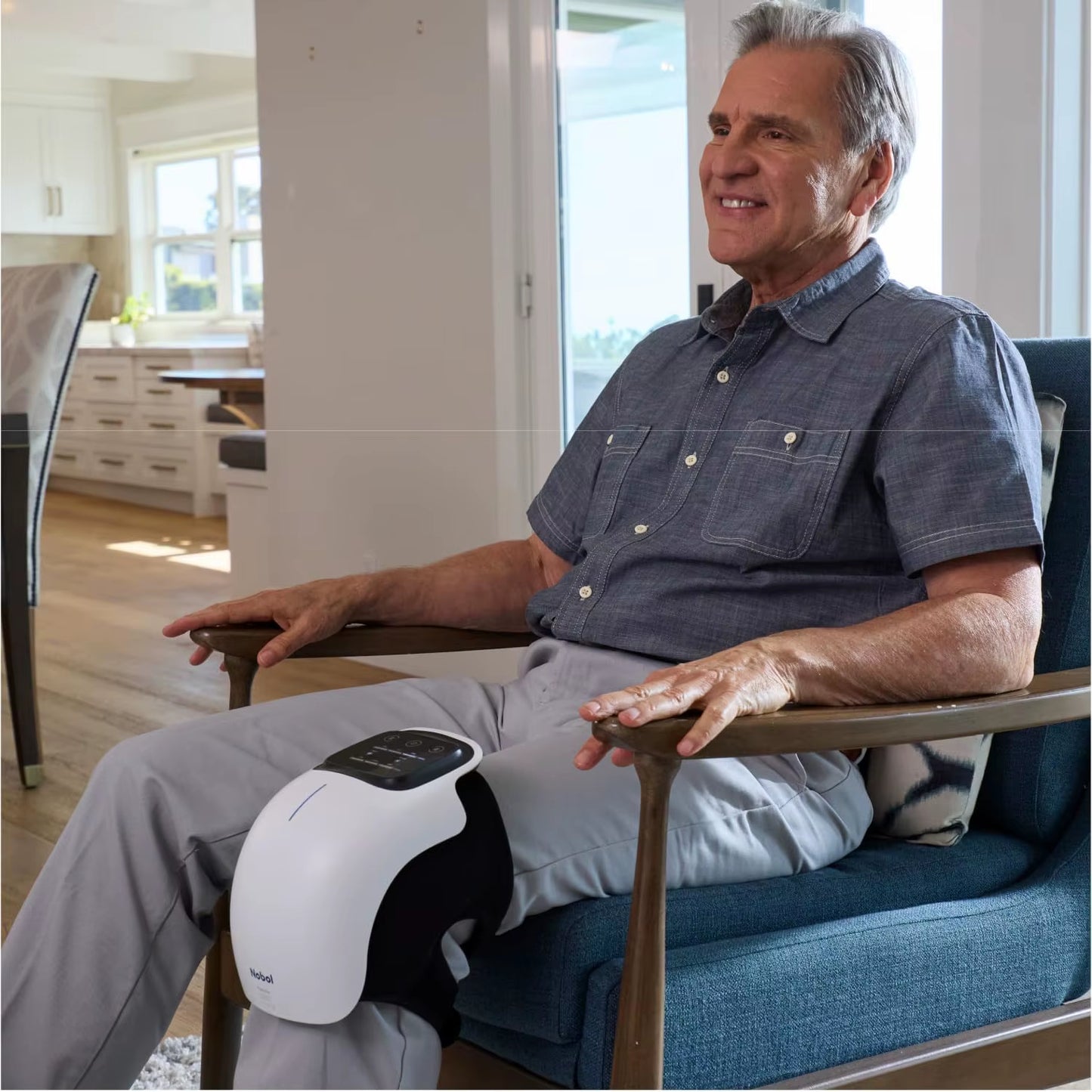 homme en chemise bleu et pantalon gris assis avec appareil massage pour le genou dans le salon