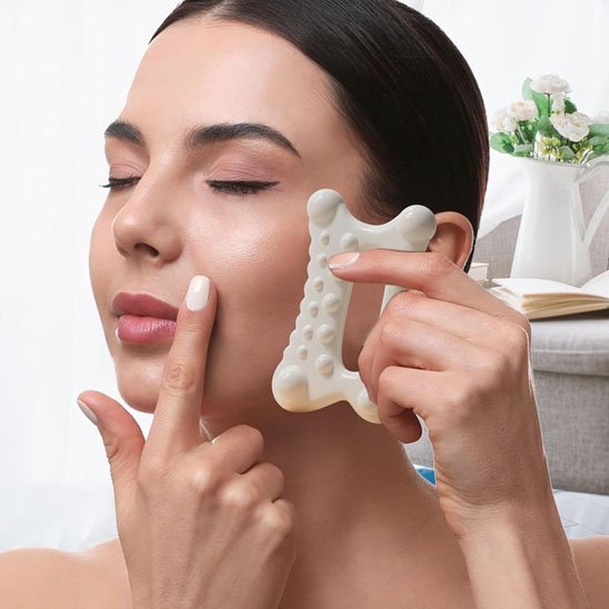 femme un ongle blanc avec un gua sha sur son visage devant un petit sofa