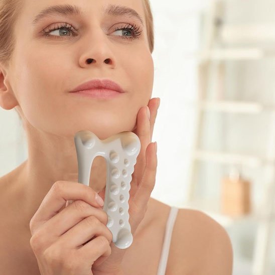 femme une main sur son visage et l'autre sur un gua sha blanc devant un étagère