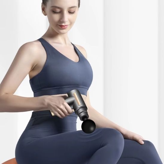 femme en camisole bleue et pantalon bleu avec pistolet de massage musculaire vert devant un rideau blanc