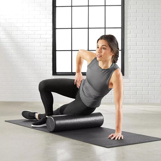 femme en pantalon noir camisole gris la cuisse sur rouleau de massage noir devant fenêtre a carreau